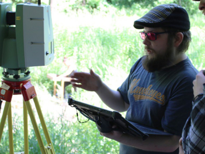 Man outside holding rugged tablet looking at surveying equipment